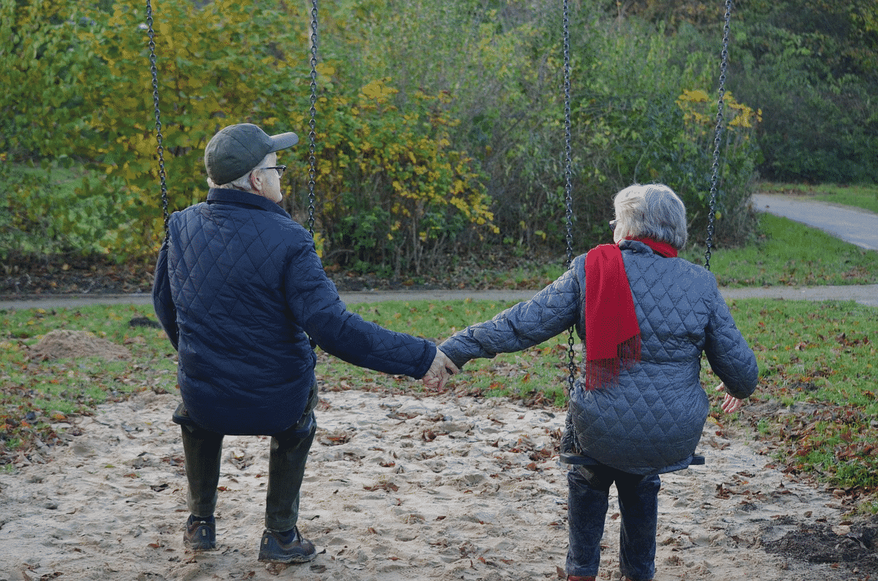 An elderly couple that is holding hands and swinging at a park.