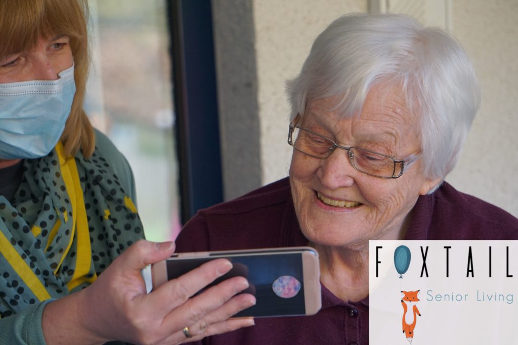 A masked assisted living facility staff member showing a resident something on a phone