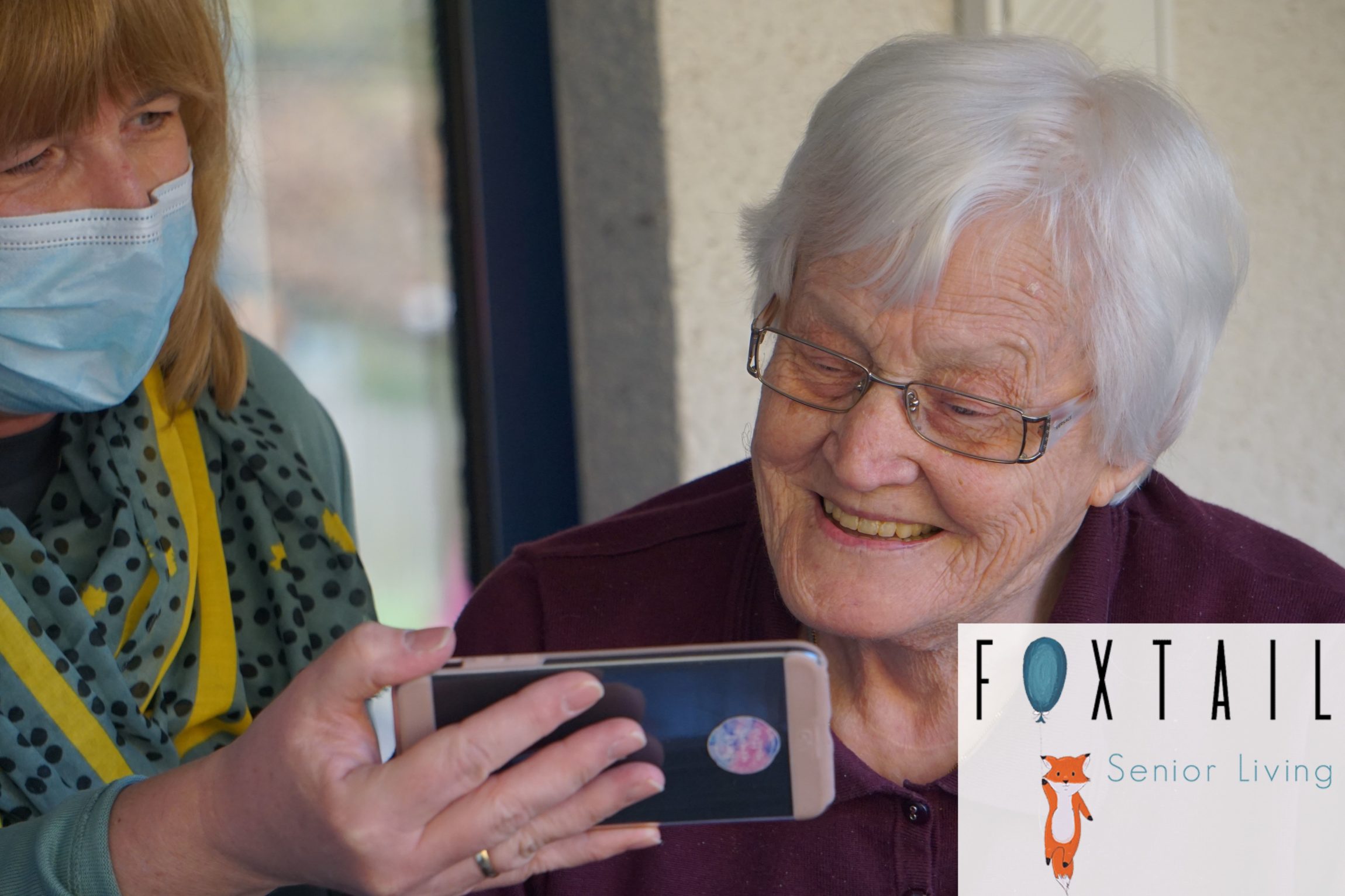 A masked assisted living facility staff member showing a resident something on a phone